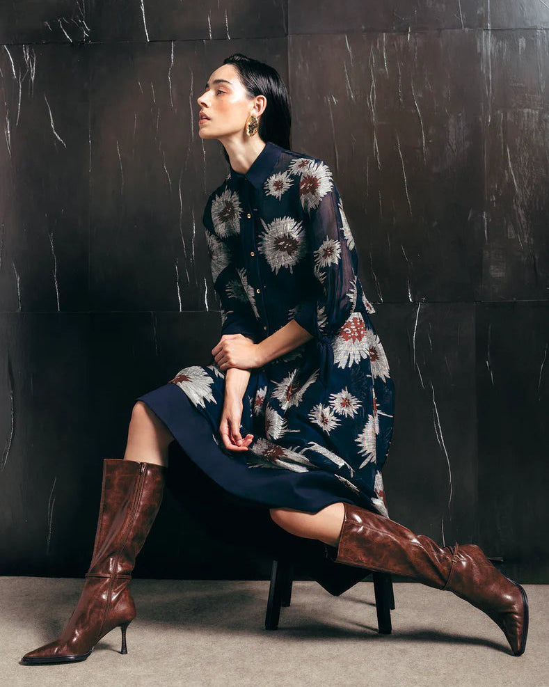 Woman in a floral dress and brown knee-high boots against a dark textured wall.